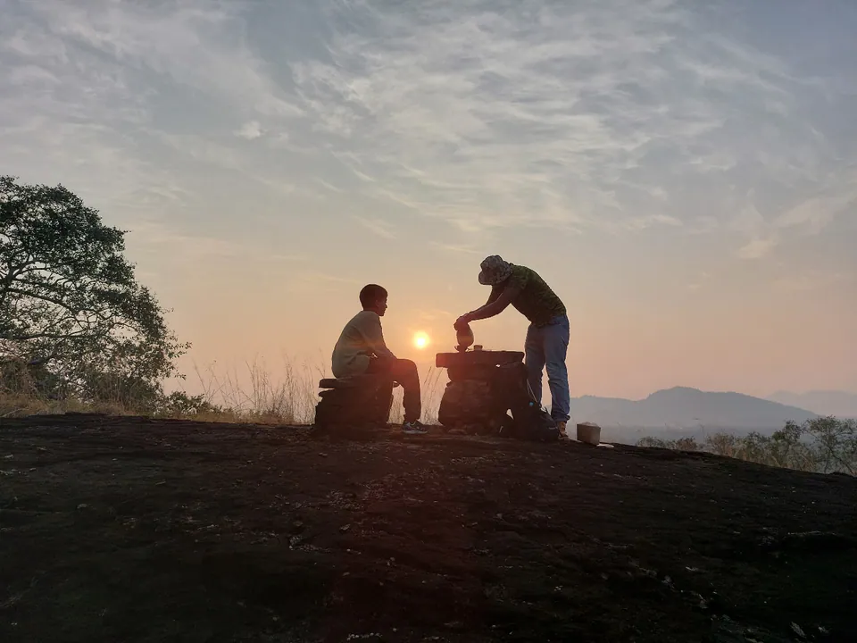 Two figures silhouetted on a rock summit at sunrise, hazy hills beyond