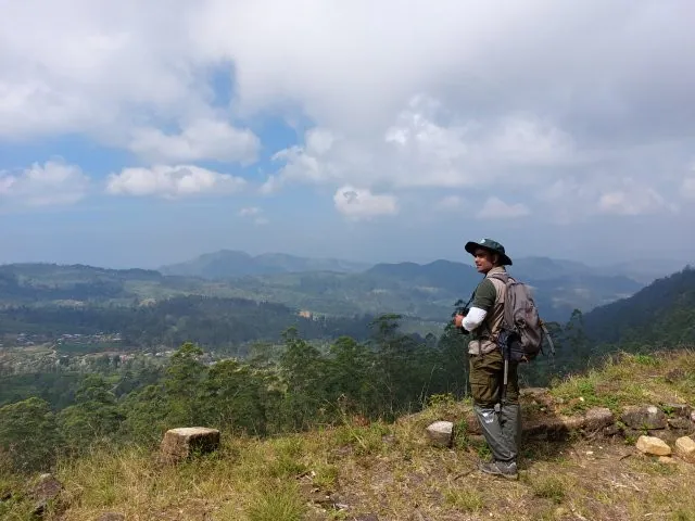 A birder in a bush hat with binoculars at a hill-country viewpoint