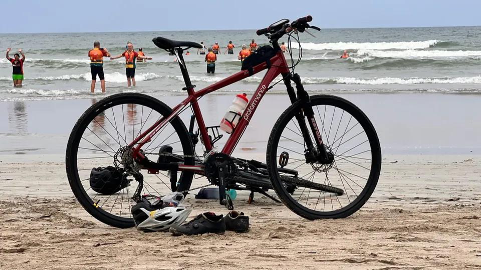 A red bike standing on the sand beside helmets and cycling shoes, as riders in orange jerseys wade into the surf