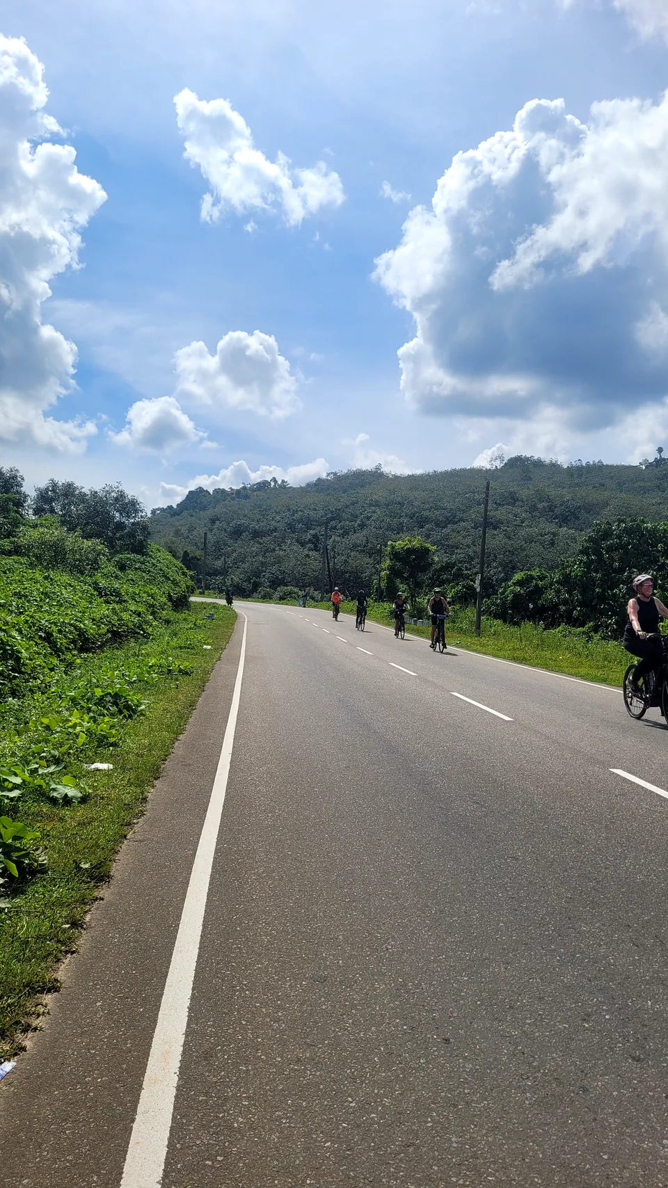 A long straight country road with a line of distant cyclists, big white clouds over forested hills
