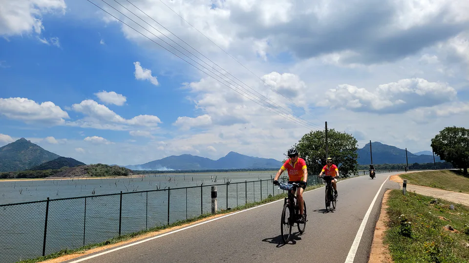 Two cyclists riding a reservoir bund road past drowned trees, with peaked mountains beyond