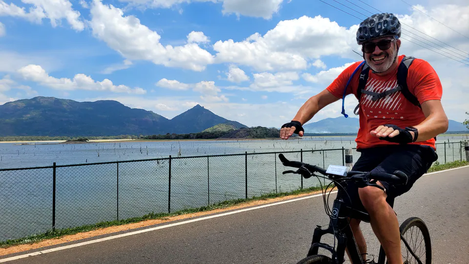 A grinning cyclist waves from his bike beside a reservoir and peaked mountains