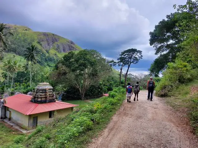 A trekker walking a village road past a small Hindu kovil beneath a rocky mountain