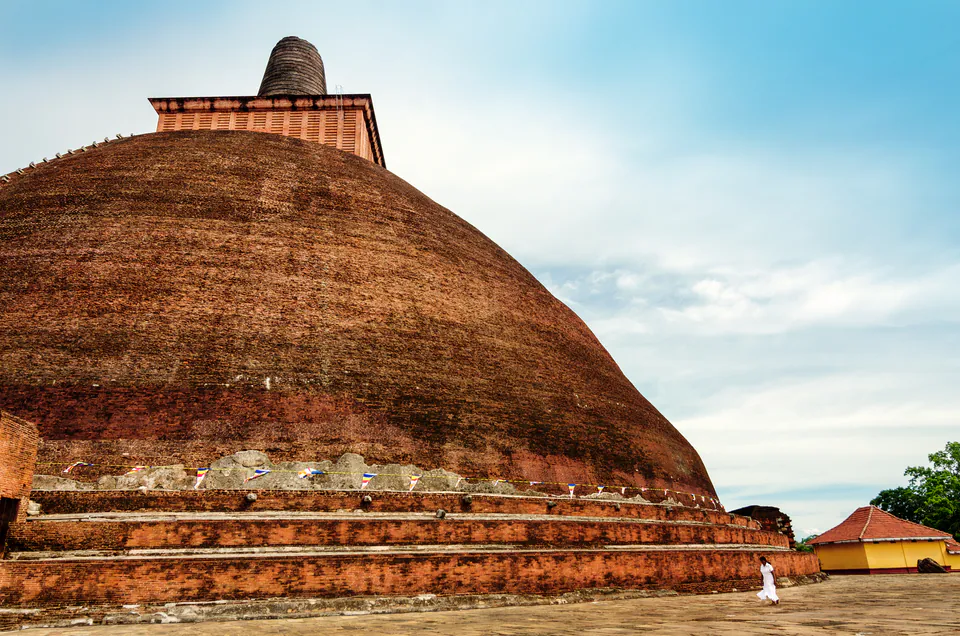 The colossal brick dome of an ancient dagoba ringed with Buddhist flags, a lone pilgrim in white crossing the terrace