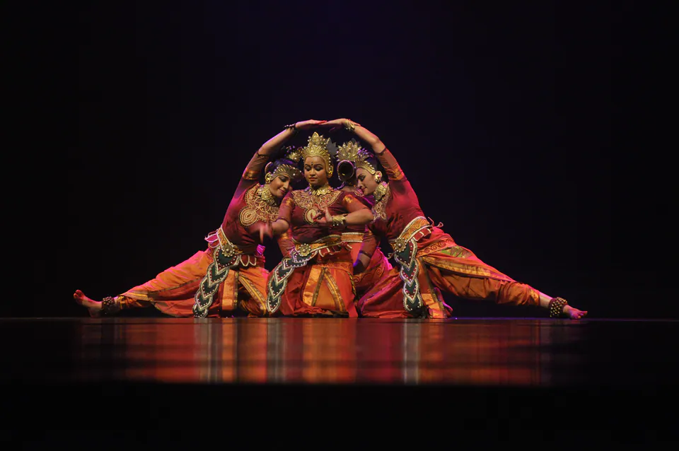 Three dancers in red and gold costumes and golden crowns forming an arch with their arms on a dark stage