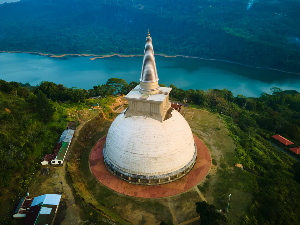 A white hilltop stupa with a tall ringed spire above a turquoise reservoir and forested hills, seen from the air
