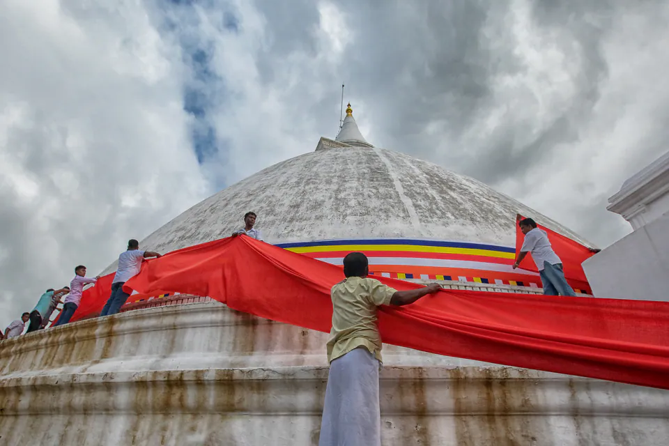 Devotees stretching a long red cloth around the dome of a white dagoba as an offering