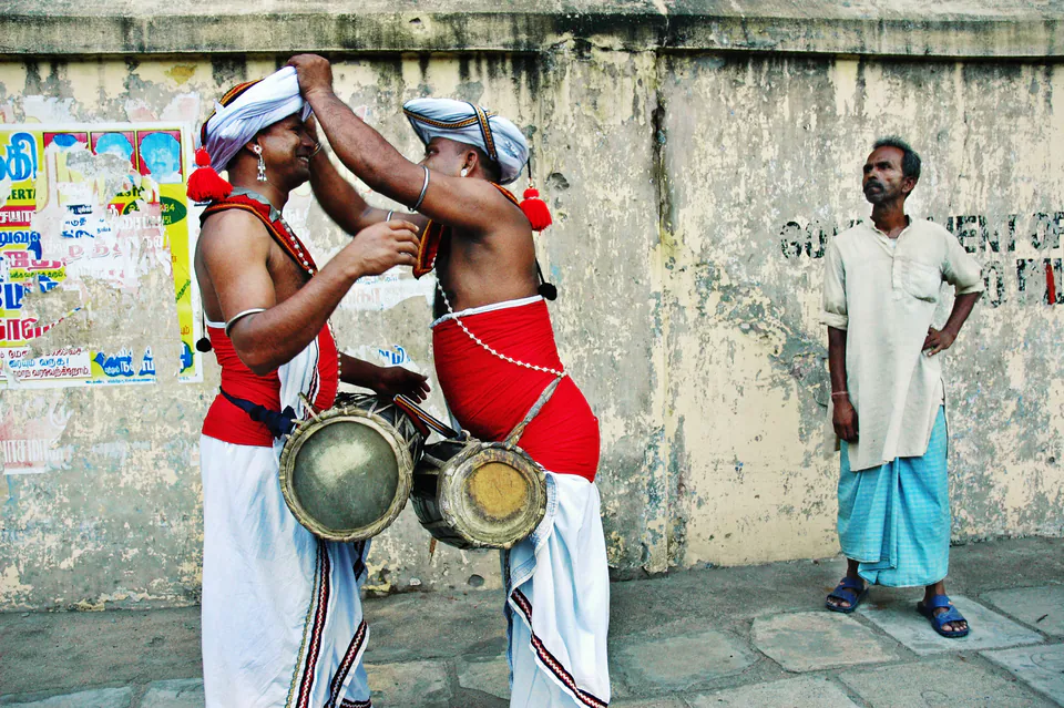 Two drummers in white turbans and red sashes adjusting each other’s headdress beside their drums