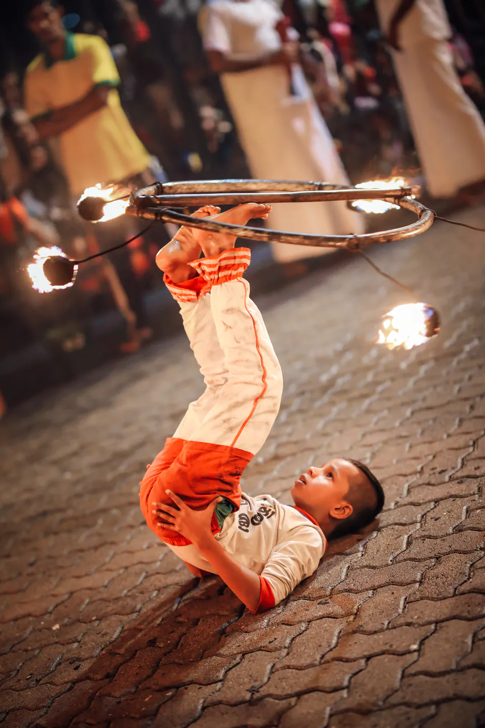 A boy lying on the paving at night, balancing a ring of flaming torches on his feet as a crowd looks on