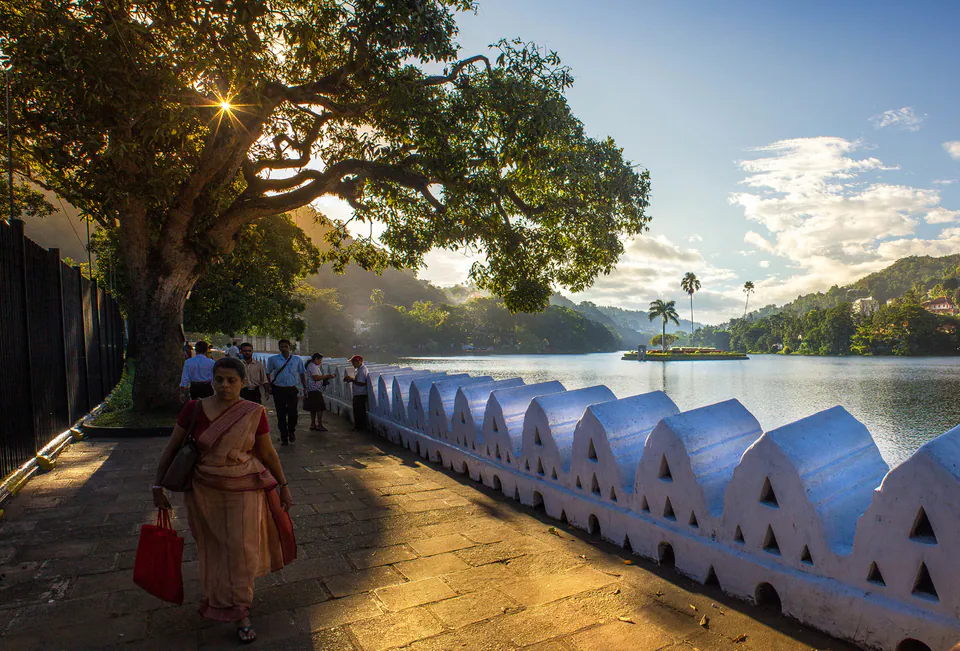 A woman in a sari walking the shaded lakeside promenade beside the white scalloped wall of Kandy Lake