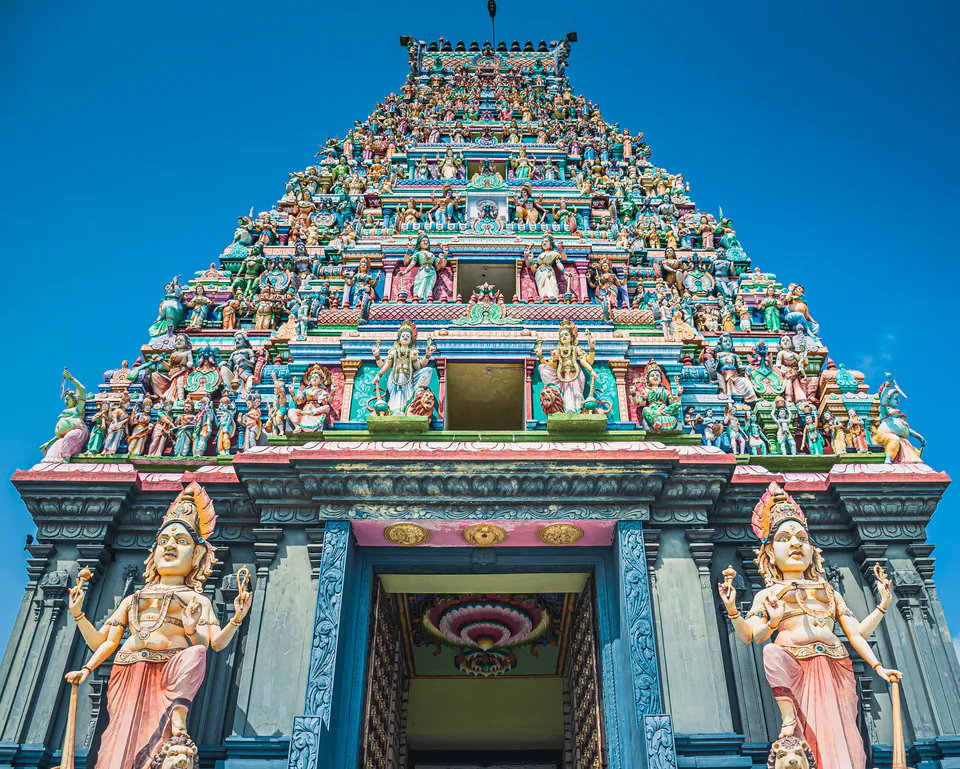 The painted gopuram of a Hindu kovil crowded with deities, two guardian statues flanking the door