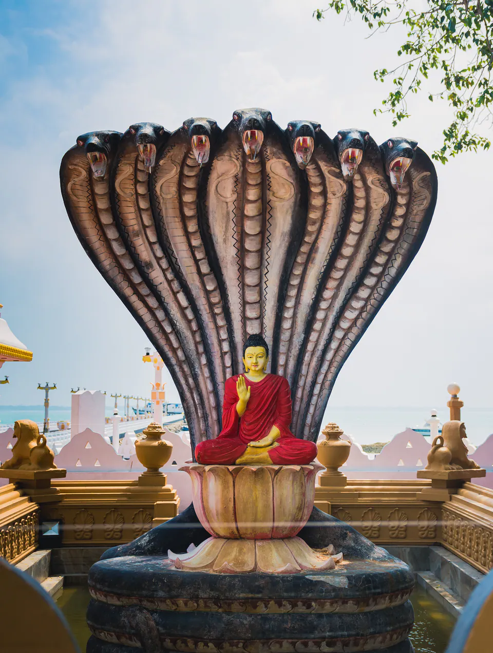 A Buddha in a red robe seated beneath a seven-headed cobra hood on a lotus plinth, the sea beyond