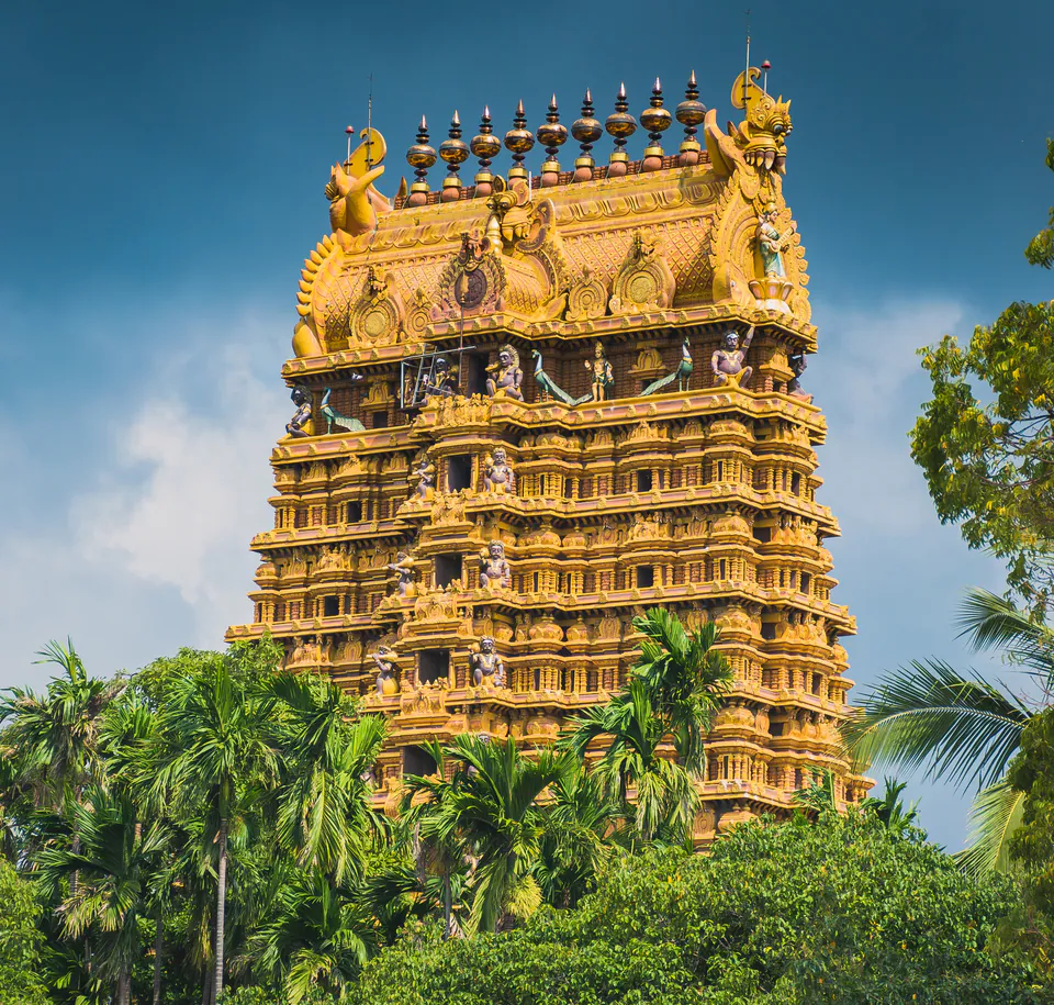 The tiered golden gopuram of a Hindu temple crowded with sculpted figures, rising above areca palms