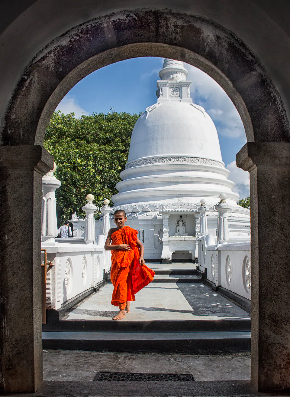A young novice monk in an orange robe walking barefoot through a stone archway, a white stupa behind