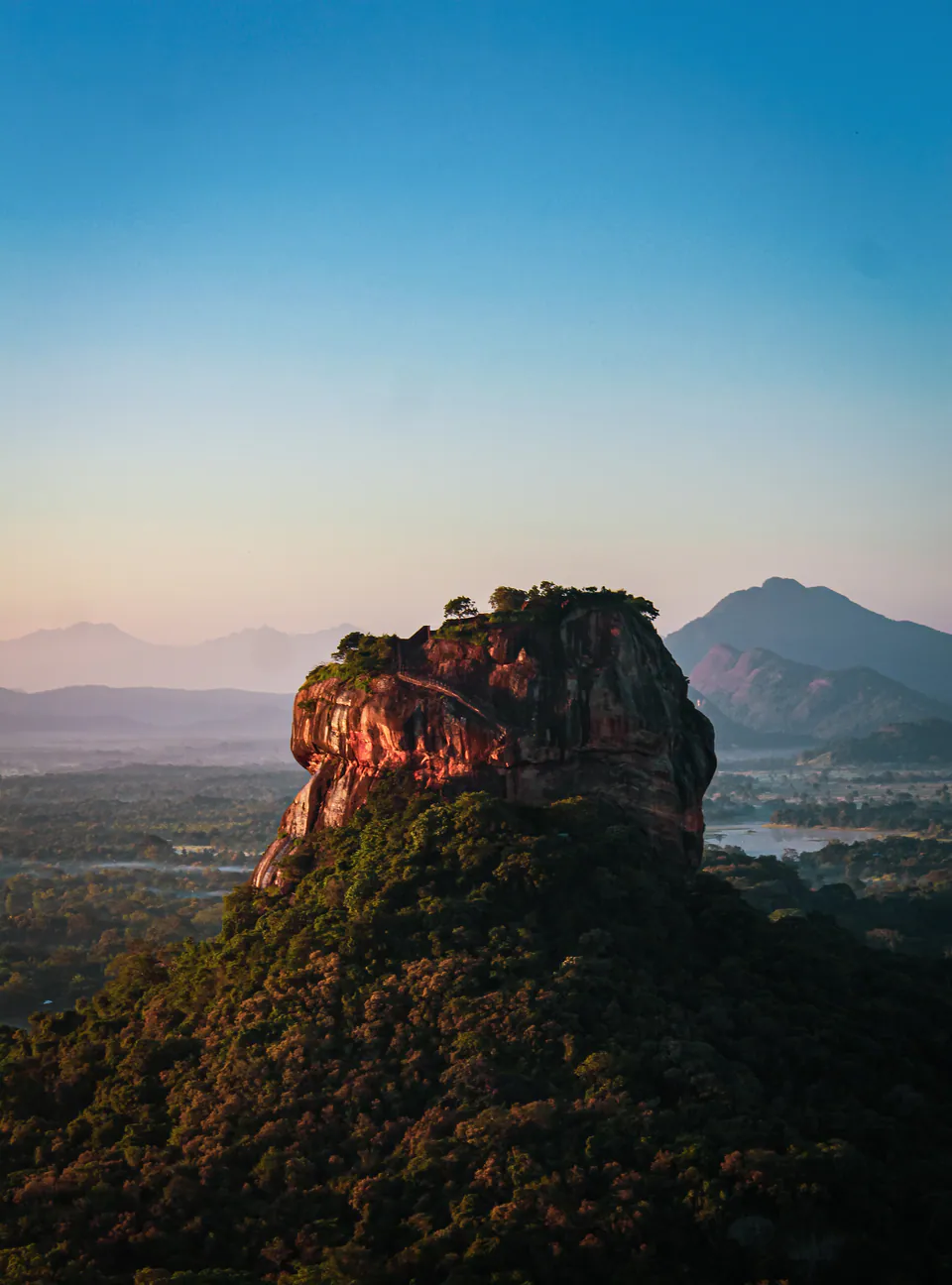 Sigiriya rock glowing red in the first light above the forest, misty plains and blue mountains beyond