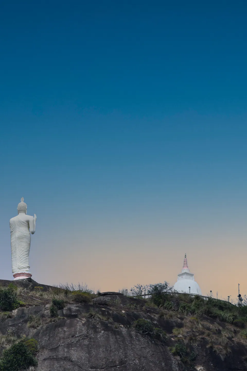 A giant white standing Buddha and a small dagoba on a rocky hilltop against a clear dusk sky