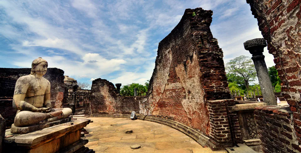 A weathered seated Buddha inside the curved brick walls of the ruined Vatadage, a carved stone pillar beside it