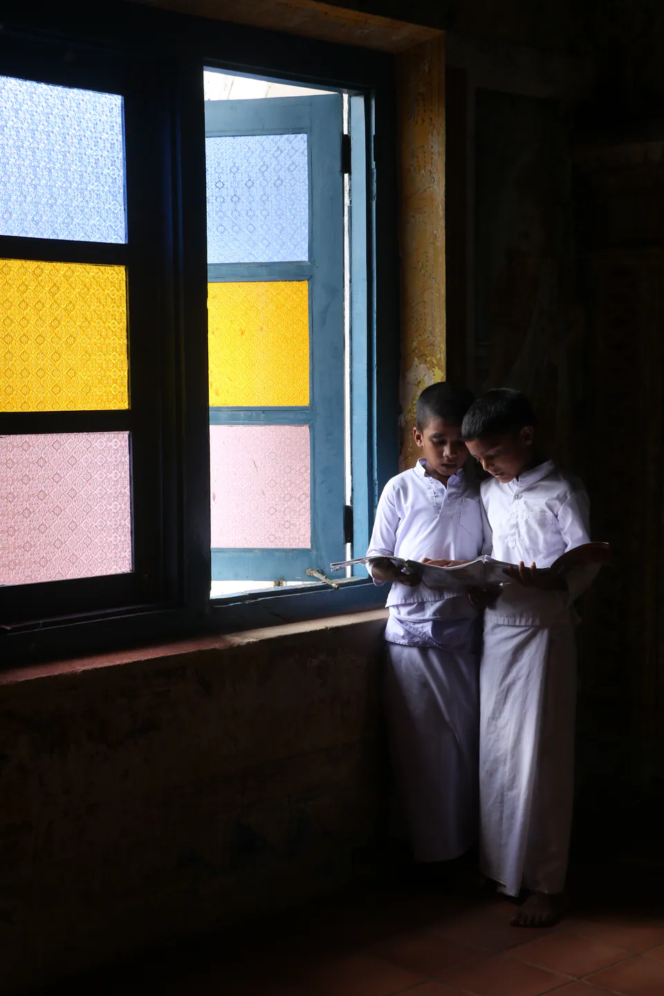 Two boys in white reading a book together by a window of coloured glass in a temple