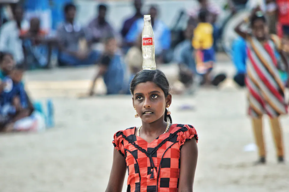 A girl in a checked dress balancing a glass bottle on her head in a race at a village games day, spectators seated behind