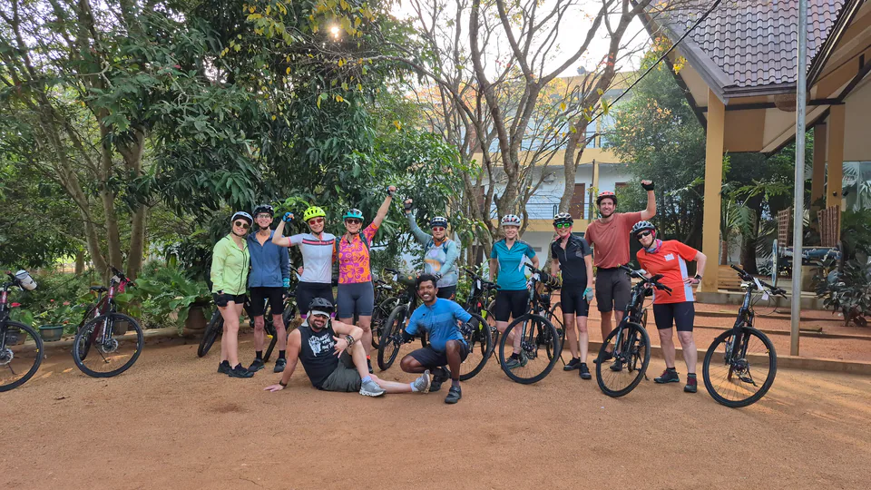 A cycling group cheering with arms raised beside their bikes in a leafy hotel garden, their guide crouching in front