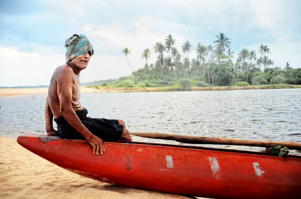 An elderly fisherman seated on his red outrigger canoe beneath the palms