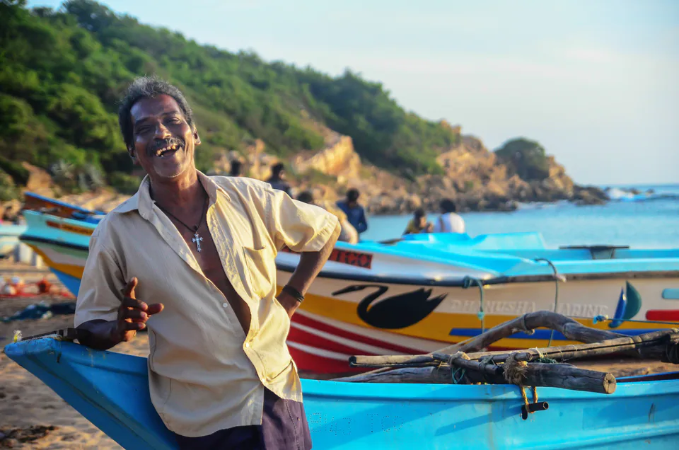 A laughing fisherman leaning on his bright blue boat on the beach below a green headland