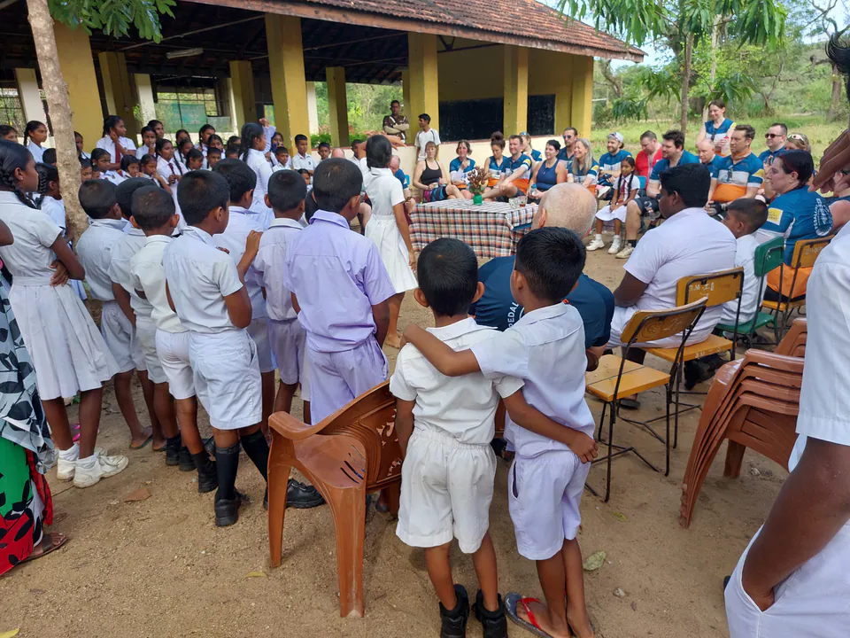 Pupils in white school uniforms crowding round a visiting group at a rural school, two boys arm in arm in front