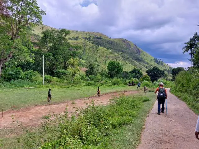 A walker passing a village field where children play cricket beneath green hills