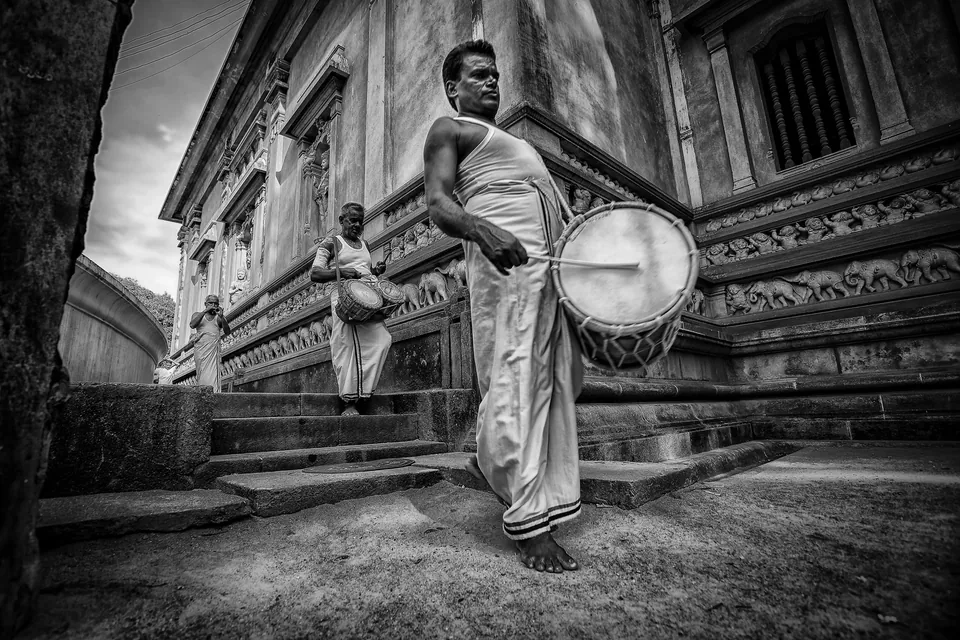 Temple drummers in white sarongs walking past a carved temple wall, in black and white