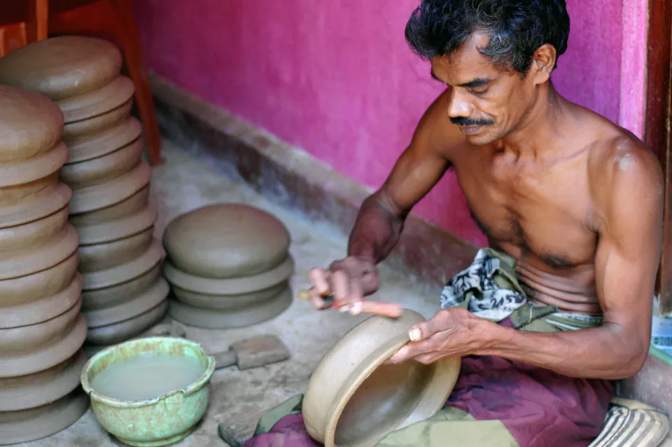 A village potter shaping a clay pot by hand beside stacks of unfired lids against a pink wall