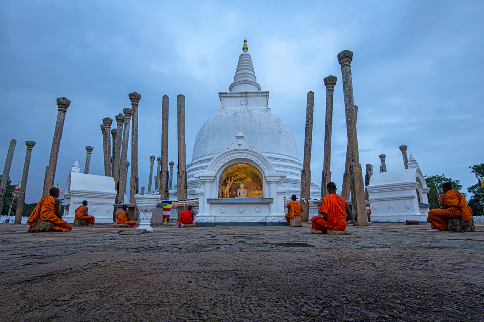 Monks seated in meditation before the white Thuparamaya dagoba and its ancient stone pillars at dusk