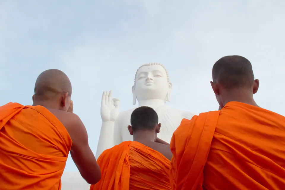 Three monks in saffron robes, seen from behind, raising their hands in worship before a giant white Buddha statue