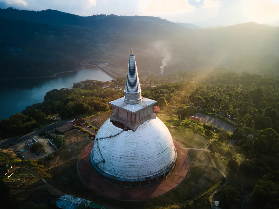 Evening light streaming over forested hills and a reservoir beside a white hilltop stupa