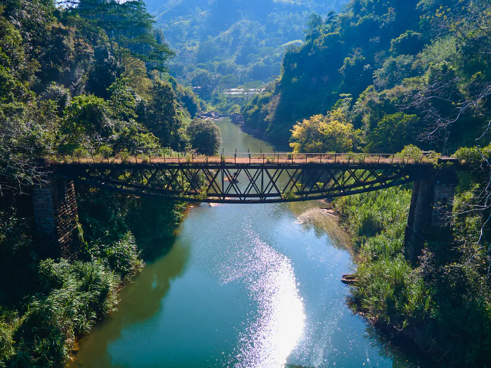 An old steel bridge spanning a river gorge between jungle-clad cliffs, seen from above