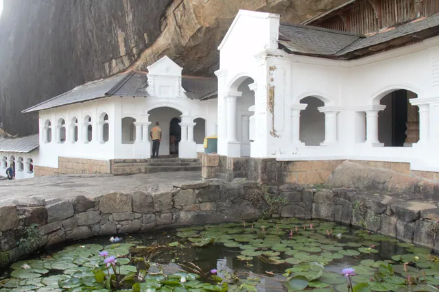 The whitewashed colonnade of the Dambulla cave temple under its overhanging rock, water lilies in front
