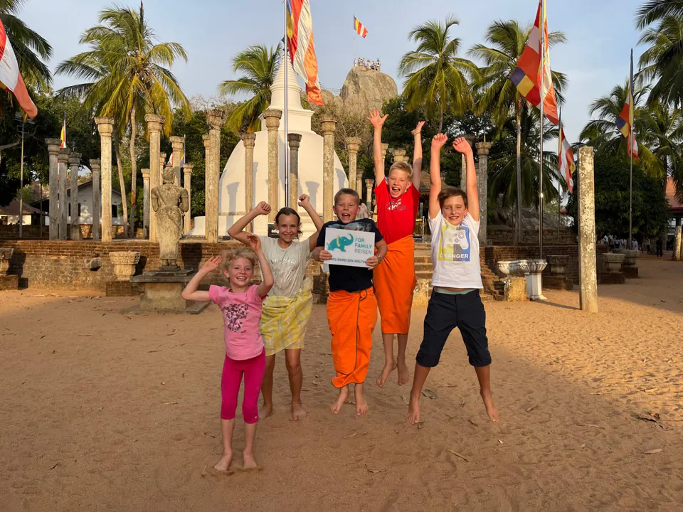 Five children in temple sarongs leaping for joy on the sand before a white dagoba ringed with ancient stone pillars
