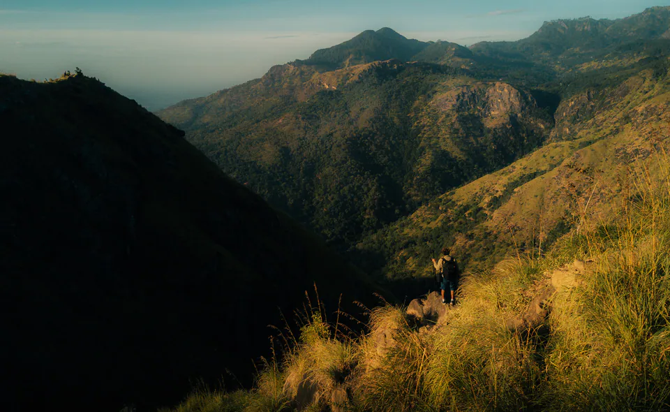 A lone hiker on a grassy ridge in golden light, with hills rolling away behind
