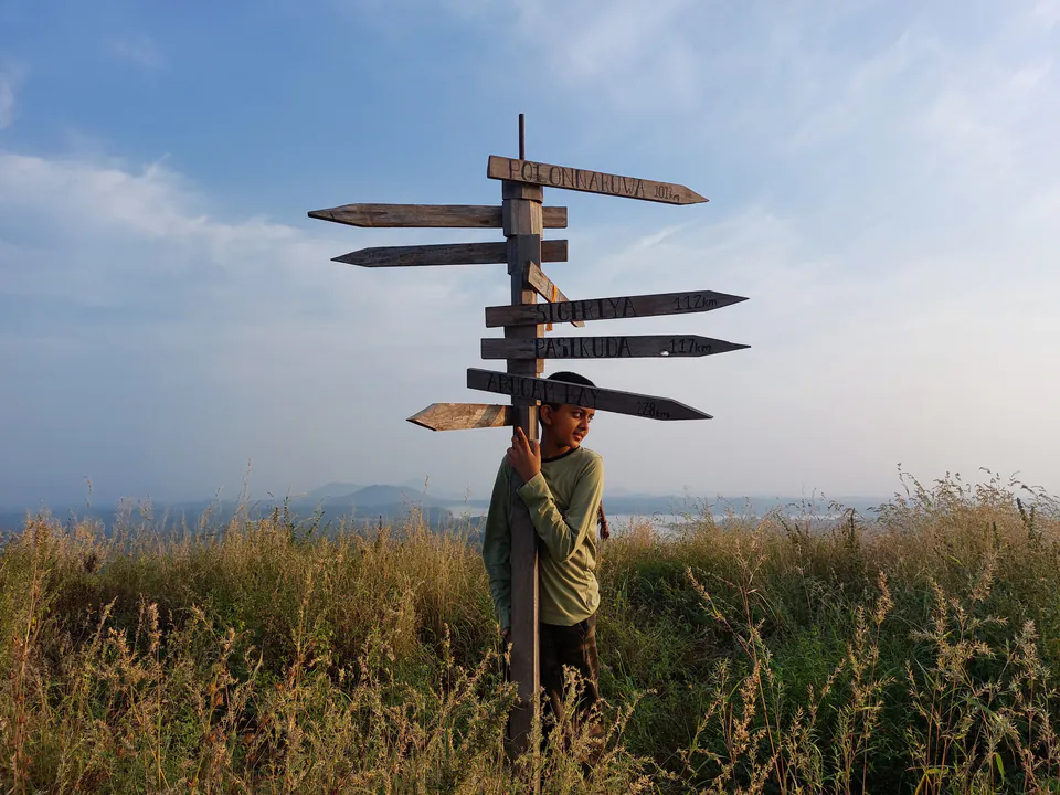 A boy hugging a wooden signpost to Polonnaruwa, Sigiriya, Pasikuda and Arugam Bay on a grassy hilltop at first light