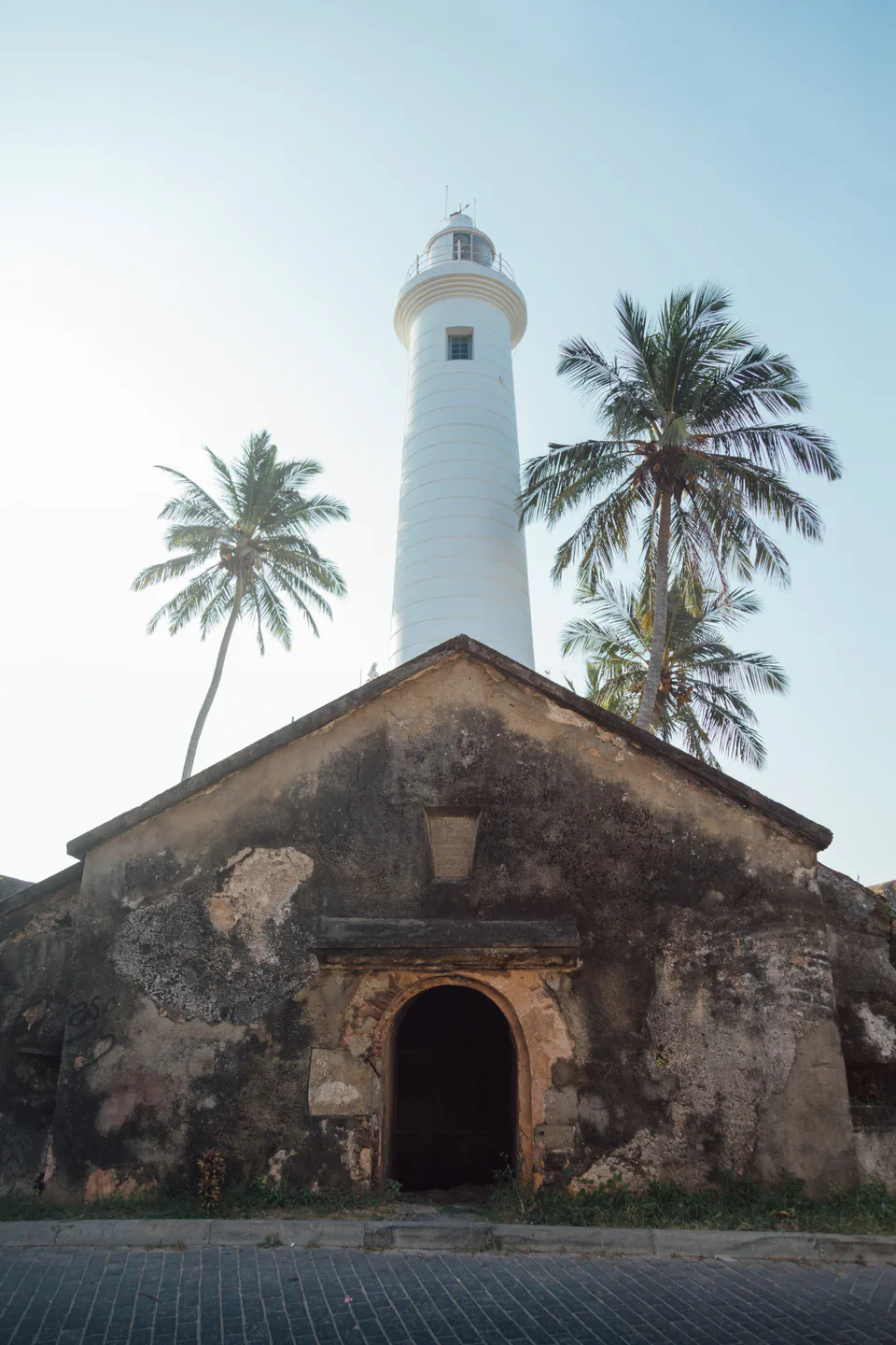 Galle lighthouse rising behind an arched Dutch-era building and coconut palms on a cobbled street