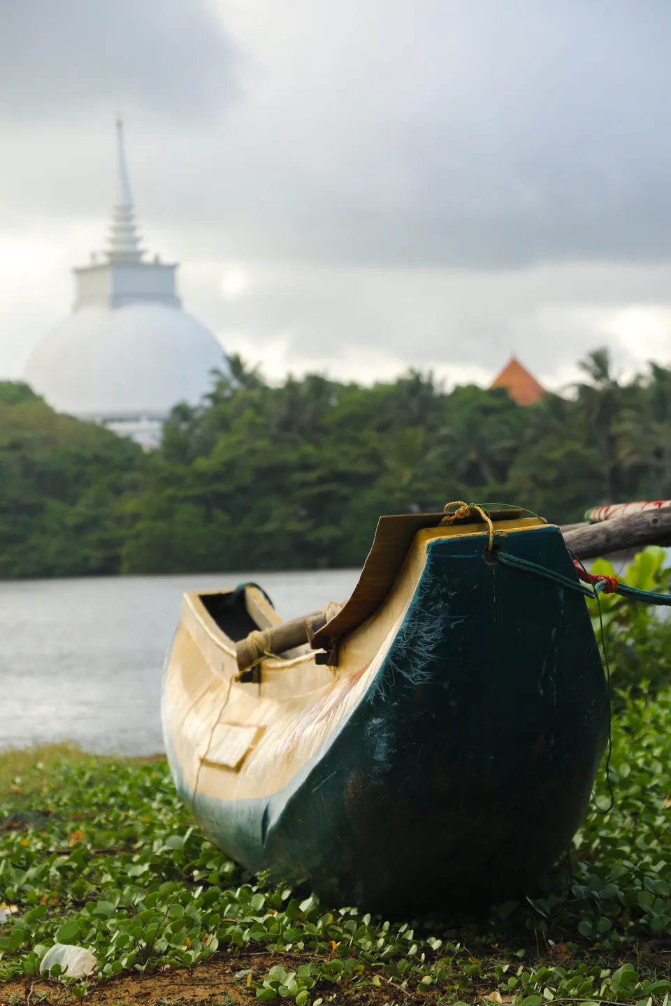 A wooden outrigger canoe drawn up on a quiet lakeshore, a white stupa beyond the trees in the mist