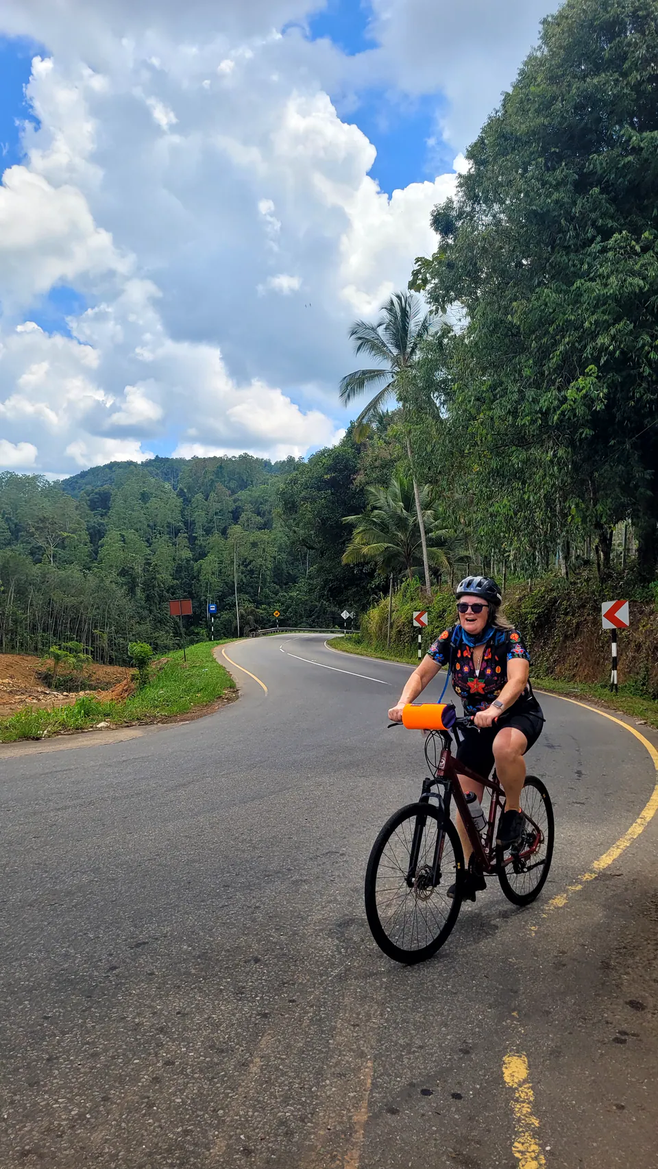 A smiling cyclist rounding a bend on a winding hill road beneath palms and towering clouds