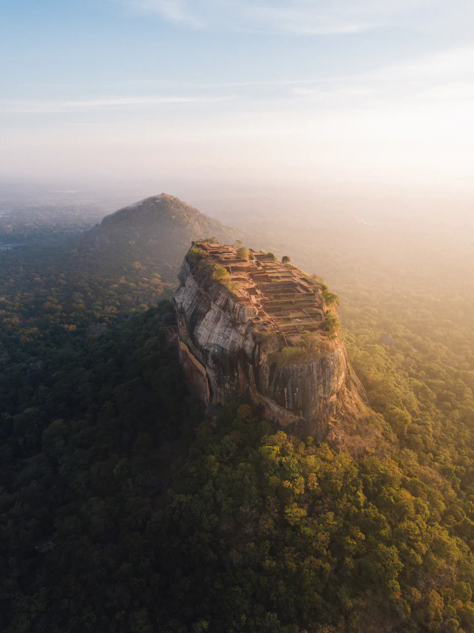 Sigiriya rock fortress and its summit ruins rising out of misty forest at golden hour, seen from the air