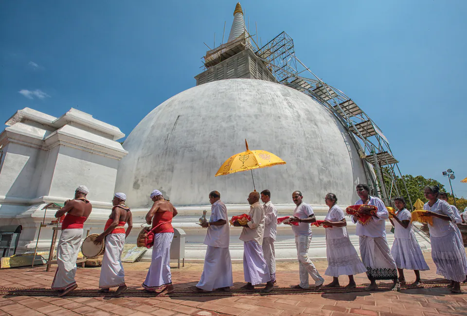 Pilgrims in white carrying offerings with drummers beneath a ceremonial umbrella, circling a white dagoba