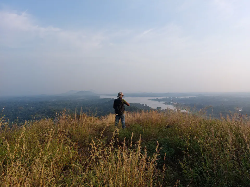 A walker in a bucket hat on a grassy hilltop at dawn, looking over misty forest and a wide reservoir