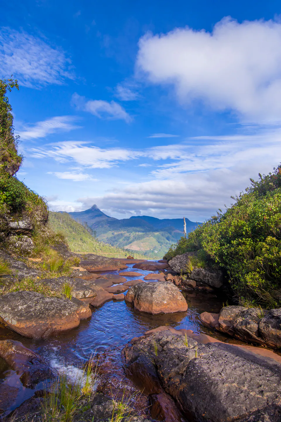 A rocky stream above a hill-country waterfall, with the pyramid of Adam’s Peak on the horizon
