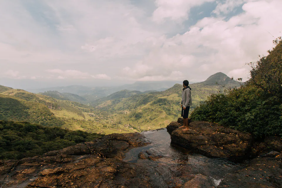 A walker on a rock ledge beside the pools at the top of Diyaluma Falls, looking over a green valley